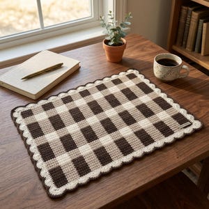 May include: A rectangular crochet placemat featuring a brown, beige, and cream checkered pattern. The placemat has a scalloped edge border in cream yarn and a small rectangular fabric tag attached to the lower right corner of the woven surface.