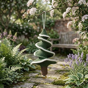 Può includere: Un ornamento all'uncinetto a forma di spirale che raffigura un albero verde scuro con bordi bianchi, un tronco marrone e una piccola campanella dorata appesa alla base. L'oggetto è dotato di un cordino bianco per essere appeso.
