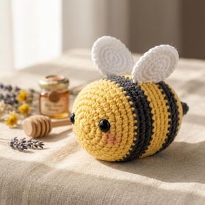 Puede incluir: Un peluche de abeja tejido a ganchillo con un cuerpo redondo de rayas horizontales alternas en amarillo y gris oscuro, dos alas blancas texturizadas en la parte superior, ojos de cuentas negras y pequeñas marcas rosas en las mejillas.