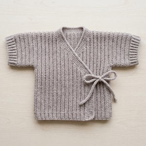 May include: A light beige, short-sleeved wrap-style cardigan featuring a vertical ribbed knit texture. The garment includes a V-neckline and a matching fabric tie closure on the side, with ribbed cuffs on the sleeves and a ribbed hem at the bottom edge.