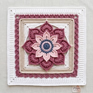 May include: A square crochet motif featuring a central dark blue circle surrounded by layers of light pink, dark pink, and beige petals. The design is framed by a decorative dark pink border and a wide white outer edge with the text Musa Stitch Studio.