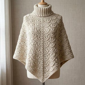 Op de afbeelding: Een beige gehaakte poncho met een hoge geribbelde col en een herhalend waaiervormig steekpatroon over het hele lichaam. Het kledingstuk wordt getoond op een paspop en heeft een puntige zoom en ingewikkeld, getextureerd haakwerk.