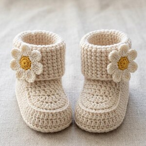 May include: A pair of cream-colored crochet baby booties featuring a folded cuff design. Each bootie is adorned with a single crocheted flower appliqué on the outer side, consisting of white petals and a textured mustard yellow center.