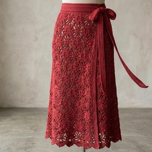 Peut inclure: Une jupe portefeuille en crochet rouge foncé présentant un motif floral complexe sur tout le tissu. La jupe possède une ceinture unie avec un long lien assorti sur le côté, formant un nœud. L'ourlet présente une bordure festonnée.