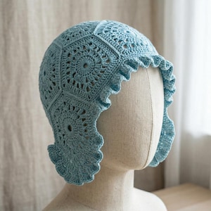 May include: A light blue crochet bonnet constructed from multiple hexagonal motifs featuring a central circular pattern. The headwear includes a ruffled edge that frames the face, providing a textured appearance with intricate yarn stitching.