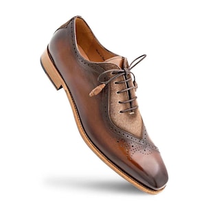 Könnte beinhalten: Ein brauner Oxford-Schuh aus Leder mit polierter Schuhspitze, aufwendigen Brogue-Verzierungen an den Nähten und einem strukturierten, bronzefarbenen Einsatz am Schnürbereich. Der Schuh hat dünne braune Schnürsenkel und einen hellen Holzabsatz.