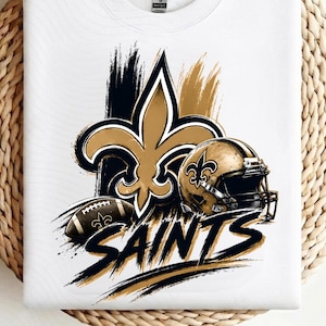 May include: A white crew neck t-shirt featuring a graphic print of a gold and black fleur-de-lis emblem, a football helmet, and a football. The word SAINTS is printed in bold, black, brush-stroke lettering below the central sports imagery.
