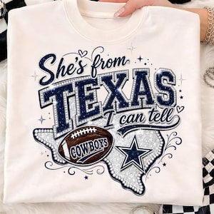May include: A cream-colored crewneck sweatshirt featuring a graphic print that reads "She's from TEXAS I can tell" in dark blue letters. The design includes a silver glitter map of Texas, a brown football labeled "COWBOYS," and a blue star.