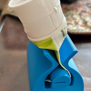 Puede incluir: Un exprimidor de tubos de pasta de dientes de plástico azul brillante con un cilindro giratorio central con una ranura. Un tubo de pasta de dientes Colgate blanco está insertado en el dispositivo, con la parte inferior pasada por la ranura.