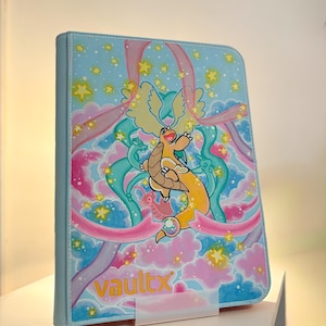 May include: A light blue trading card binder featuring a central orange dragon-like creature surrounded by pink clouds, teal ribbons, and yellow stars. The word vaultx is printed in orange at the bottom left corner of the front cover.