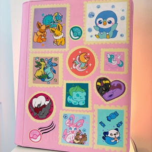 May include: A light pink binder featuring a collection of colorful, stamp-shaped illustrations of various cartoon creatures. The designs include small characters in different poses, a heart-shaped frame, and a circular emblem with a pink pokeball icon.