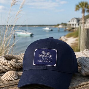 Puede incluir: Una gorra de béisbol azul marino con un parche rectangular en la parte delantera. El parche muestra un gráfico bordado en blanco de una rama de pino con una piña sobre olas estilizadas, con el texto TIDE & PINE debajo del gráfico.