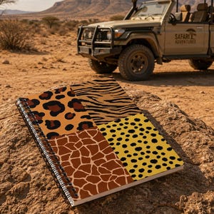 May include: A spiral-bound notebook featuring a cover divided into four distinct animal print sections: leopard spots, zebra stripes, giraffe patterns, and cheetah spots, resting on a large rock in a dry, arid landscape.