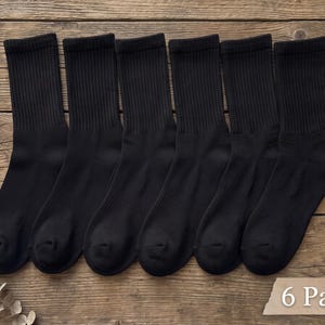 May include: A set of six pairs of solid black crew socks featuring a ribbed texture on the upper portion. The socks are arranged side by side, and a small rectangular label in the bottom right corner displays the text 6 Pairs.