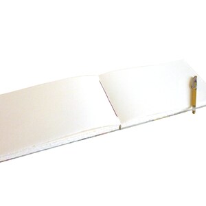 May include: An open landscape-oriented sketchbook with blank white pages lying flat. A slim, clear-barreled pen with a gold-colored cap and tip rests diagonally across the right-hand page of the notebook, which features a visible spine binding.