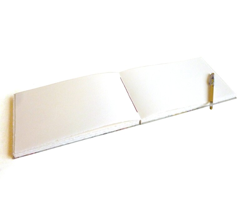 May include: An open landscape-oriented sketchbook with blank white pages lying flat. A slim, clear-barreled pen with a gold-colored cap and tip rests diagonally across the right-hand page of the notebook, which features a visible spine binding.