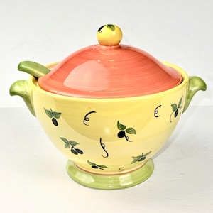 May include: A ceramic soup tureen with a yellow body featuring painted black olive motifs and green leaves. It includes a matching orange lid with a yellow knob, two side handles, and a green base. A small ceramic ladle rests inside the bowl.