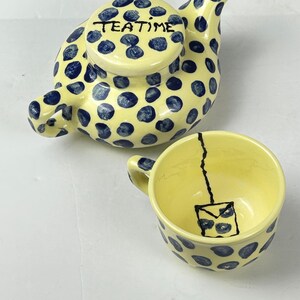 May include: A yellow ceramic teapot and matching teacup decorated with dark blue polka dots. The teapot lid features the hand-painted black text "TEATIME". The interior of the teacup displays a black line drawing of a tea bag hanging from the rim.
