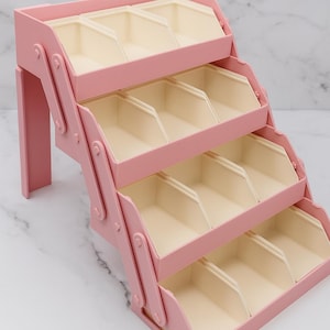 May include: A tiered storage organizer featuring a pink frame with four levels of open, cream-colored bins. The structure is supported by side panels with a hinged, expandable design, creating a staircase-like arrangement for organizing small items.