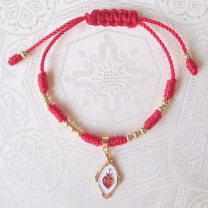 May include: A red cord bracelet featuring a gold-toned metal charm with a white enamel inlay depicting a red Sacred Heart symbol. The bracelet includes small gold-toned spherical beads and a sliding knot closure, with additional red cord ends finished with gold-toned beads.
