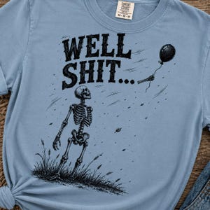 May include: A light blue t-shirt featuring a black graphic of a skeleton standing in grass, looking up at a black balloon floating away. The text WELL SHIT... is printed in black above the skeleton. The shirt has a crew neck and short sleeves.