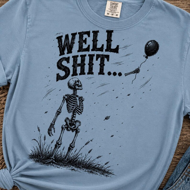 May include: A light blue t-shirt featuring a black graphic of a skeleton standing in grass, looking up at a black balloon floating away. The text WELL SHIT... is printed in black above the skeleton. The shirt has a crew neck and short sleeves.