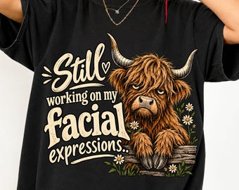 May include: A black t-shirt featuring a graphic of a brown Highland cow with long horns resting its hooves on a wooden fence. The text reads Still working on my facial expressions. Small white daisy flowers are positioned around the cow.