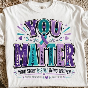 Op de afbeelding: Een wit t-shirt met een opdruk met de tekst YOU MATTER in grote paarse en turquoise letters. Een turquoise en paars bewustzijnslint staat in het midden. De tekst bevat LOVED, HEARD, SEEN, WORTHY, ENOUGH, VALUED en YOUR STORY IS STILL BEING WRITTEN.