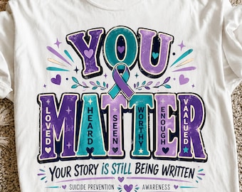 May include: A white t-shirt featuring a graphic with the text YOU MATTER in large purple and teal letters. A teal and purple awareness ribbon sits in the center. Smaller text includes LOVED, HEARD, SEEN, WORTHY, ENOUGH, VALUED, and YOUR STORY IS STILL BEING WRITTEN.