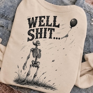 May include: A beige sweatshirt featuring a black graphic illustration of a skeleton standing in a field of grass. The skeleton holds a single black balloon. Above the skeleton, the text reads WELL SHIT... in a bold, distressed black font.