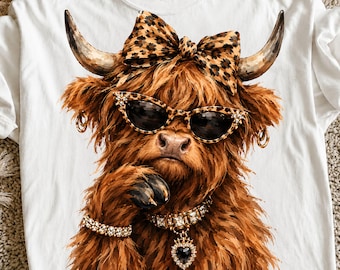 May include: A white t-shirt featuring a graphic of a brown Highland cow wearing leopard print sunglasses and a matching bow. The cow has gold hoop earrings, a crystal-encrusted collar with a heart-shaped pendant, and a matching crystal bracelet.