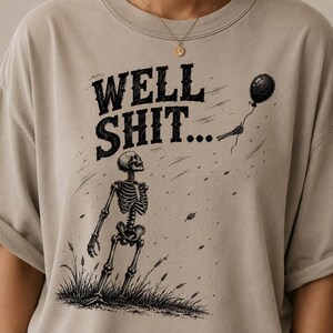 May include: A beige t-shirt featuring a black graphic of a skeleton standing in grass, looking up at a floating balloon held by a detached skeletal hand. The text "WELL SHIT..." is printed in a bold, distressed black font above the skeleton.
