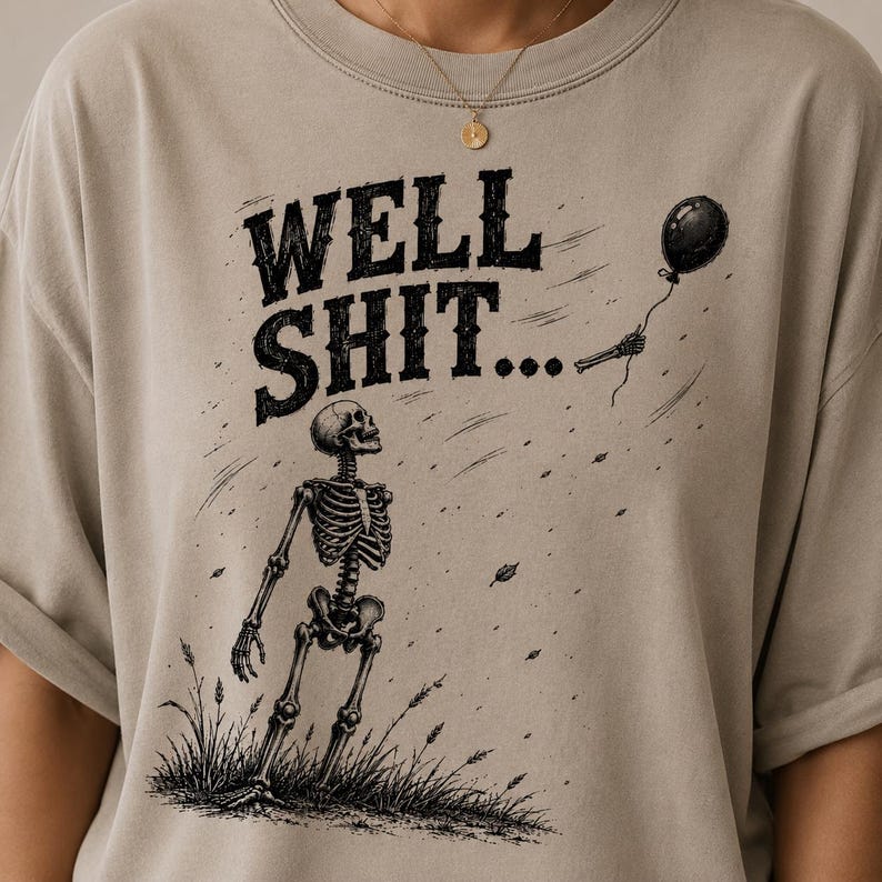 May include: A beige t-shirt featuring a black graphic of a skeleton standing in grass, looking up at a floating balloon held by a detached skeletal hand. The text "WELL SHIT..." is printed in a bold, distressed black font above the skeleton.