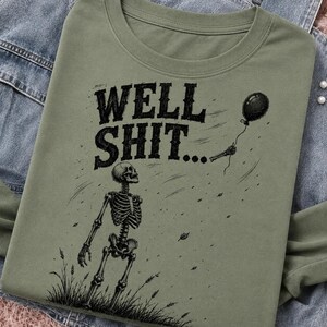 May include: An olive green t-shirt featuring a black graphic print of a skeleton standing in grass, looking up at a floating black balloon. The text "WELL SHIT..." is printed in a bold, distressed black font above the skeleton illustration.