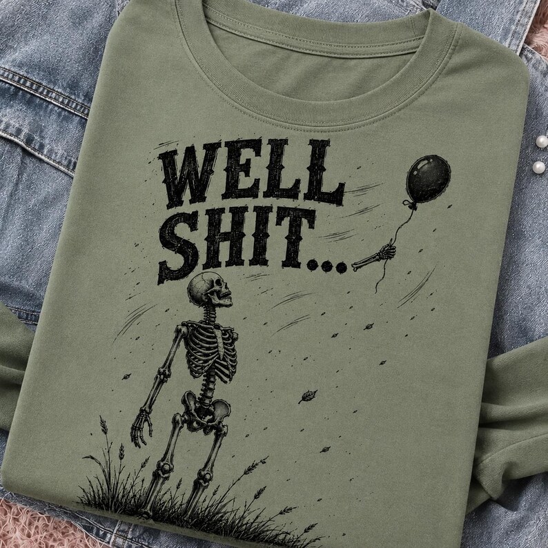 May include: An olive green t-shirt featuring a black graphic print of a skeleton standing in grass, looking up at a floating black balloon. The text "WELL SHIT..." is printed in a bold, distressed black font above the skeleton illustration.