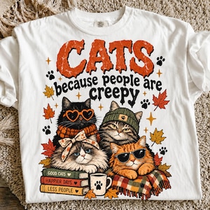 May include: A white t-shirt featuring a graphic of four cats with autumn-themed accessories, including heart-shaped glasses, a beanie, and a scarf. The text reads CATS because people are creepy, with a stack of books labelled Good Cats, Happier Days, Less People.