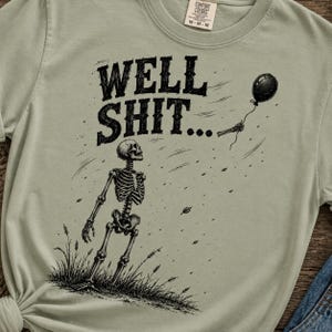 May include: A sage green t-shirt featuring a black graphic of a skeleton standing in grass, looking up at a black balloon floating away. The text WELL SHIT... is printed in a bold, distressed black font above the skeleton. The shirt has a crew neck.