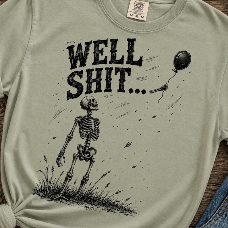 May include: A sage green t-shirt featuring a black graphic of a skeleton standing in grass, looking up at a black balloon floating away. The text WELL SHIT... is printed in a bold, distressed black font above the skeleton. The shirt has a crew neck.
