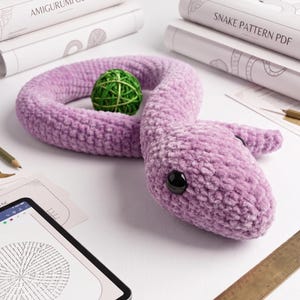 May include: A plush lavender snake toy with a textured, soft surface and black plastic eyes. The snake is coiled around a small green yarn ball. Nearby are books titled Amigurumi Guide and Snake Pattern PDF, a tablet, and a wooden ruler.