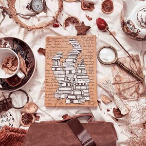 以下が含まれることがあります： 白い背景に、茶色の革のジャーナルと、家型の積み重ねられた本のイラストが描かれています。イラストは、読書と本に関する引用で囲まれています。ジャーナルは、ドライフラワー、葉、その他の自然な要素で囲まれています。