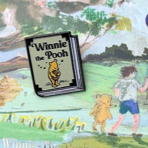 Op de afbeelding: Een emaille pin met een boek met de titel 'Winnie the Pooh'. De pin is geplaatst tegen een geschilderde achtergrond van een scène uit het verhaal, met Pooh en Christopher Robin, met een blauwe ballon.