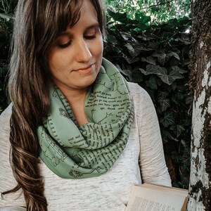 May include: A light green infinity scarf with black text and illustrations. The scarf is wrapped around the neck. The person is holding an open book with pages and a small plant. The background includes a tree and green foliage.