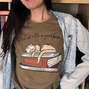 Puede incluir: Camiseta verde oliva con una ilustraci&oacute;n de un gato durmiendo sobre libros y el texto "Curl up with a good book". La persona lleva una chaqueta vaquera azul claro y una falda vaquera. Estanter&iacute;as con libros en el fondo.