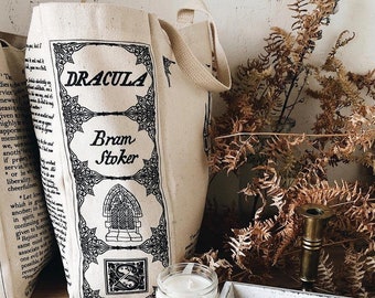 Op de afbeelding: Een witte canvas draagtas met zwarte tekst en illustraties. De tekst luidt "Dracula" en "Bram Stoker". De tas ligt op een houten oppervlak met gedroogde varens en een witte kaars in een glazen pot.
