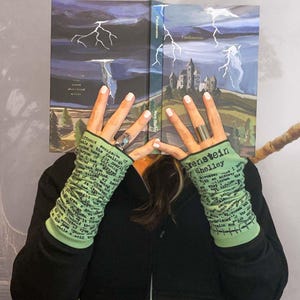 Puede incluir: Un libro de tapa dura titulado "Frankenstein" de Mary Shelley, con una ilustraci&oacute;n dram&aacute;tica de un castillo y rel&aacute;mpagos. La imagen tambi&eacute;n presenta calentadores de brazo verdes con texto y una sudadera con capucha negra.