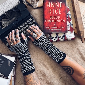 Puede incluir: Un par de guantes sin dedos negros con texto blanco impreso en ellos. Los guantes son usados por una persona con tatuajes en los brazos. La persona está escribiendo en una máquina de escribir vintage. Un libro con una cubierta roja y un diseño de calavera es visible en el fondo.