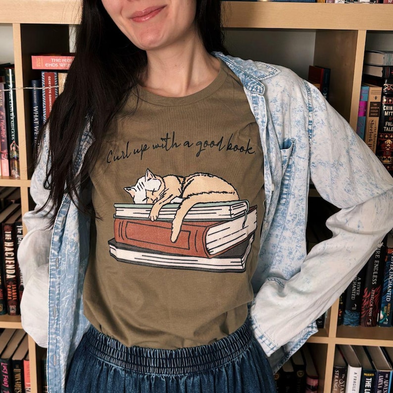 Puede incluir: Camiseta verde oliva con una ilustraci&oacute;n de un gato durmiendo sobre libros y el texto "Curl up with a good book". La persona lleva una chaqueta vaquera azul claro y una falda vaquera. Estanter&iacute;as con libros en el fondo.