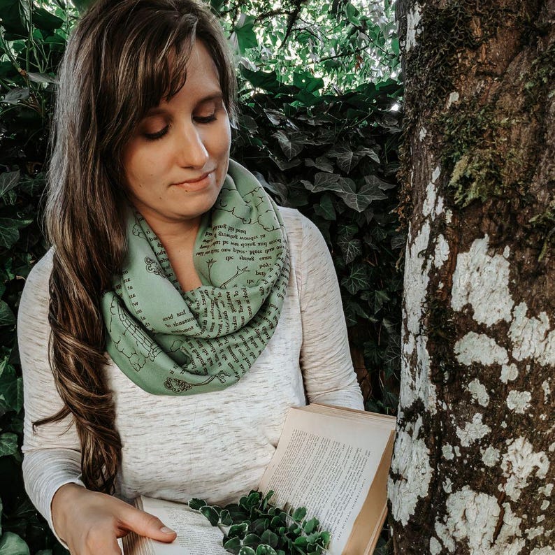 May include: A light green infinity scarf with black text and illustrations. The scarf is wrapped around the neck. The person is holding an open book with pages and a small plant. The background includes a tree and green foliage.