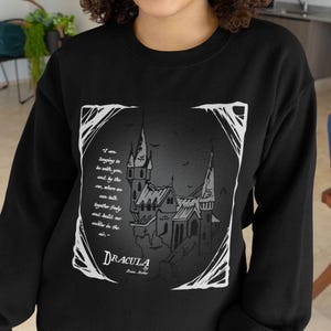 May include: Black sweatshirt featuring a white graphic print of a gothic castle and a quote from Dracula: "I am longing to be with you, and by the car, where we can talk together freely and build our castle in the air."