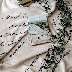 Op de afbeelding: Een beige deken met zwarte tekst die "Miss A. E. I can listen within my reach that I am most undeviating go, uncertain my fate possible when you never." luidt. Een gesloten boek met een kleurrijke illustratie van een strandscène ligt op de deken. Een groene, bladerrijke wijnstok is over de deken gelegd.